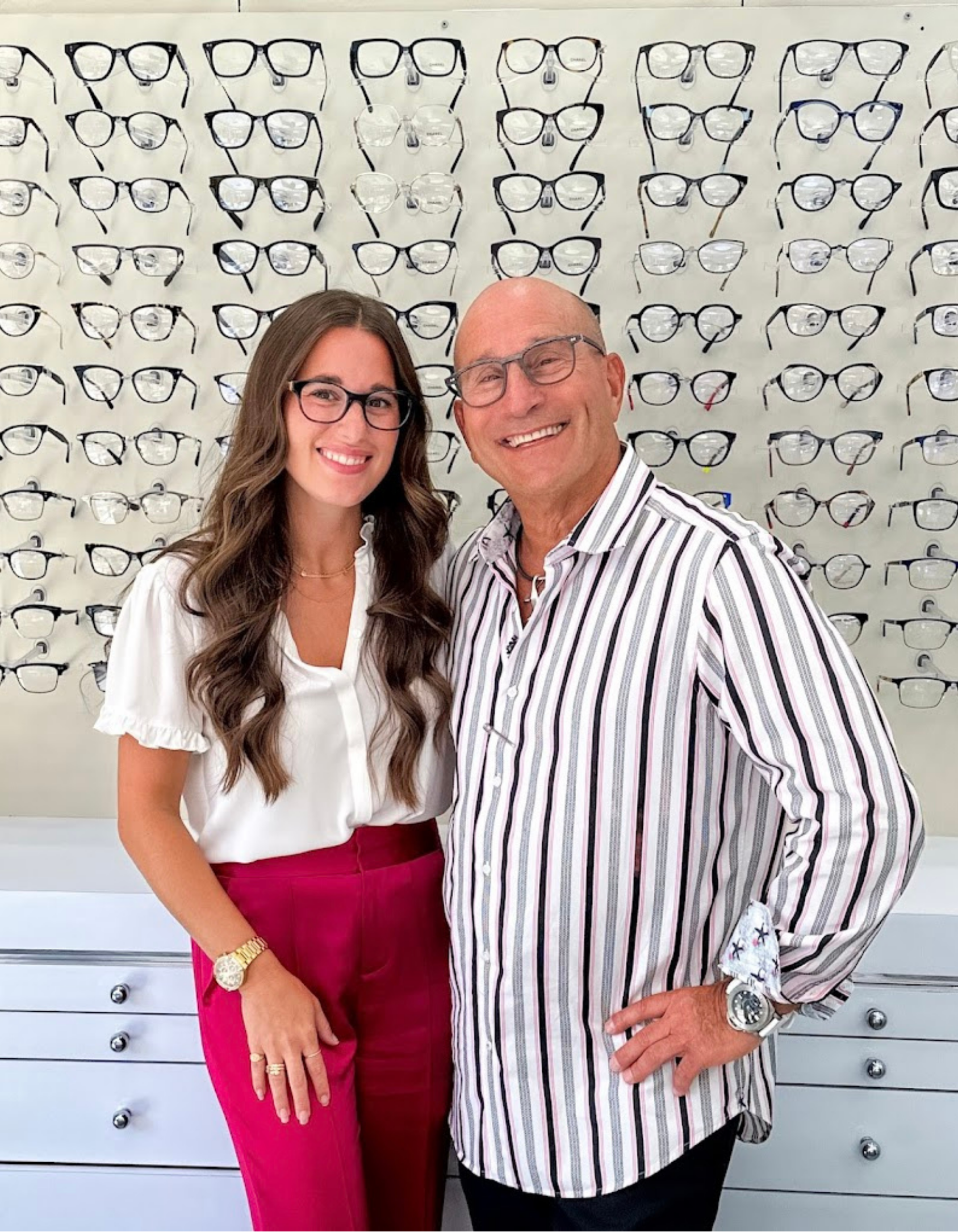 Erica and Philip Meltzer wearing glasses and smiling in front of a display wall filled with various eyeglass frames