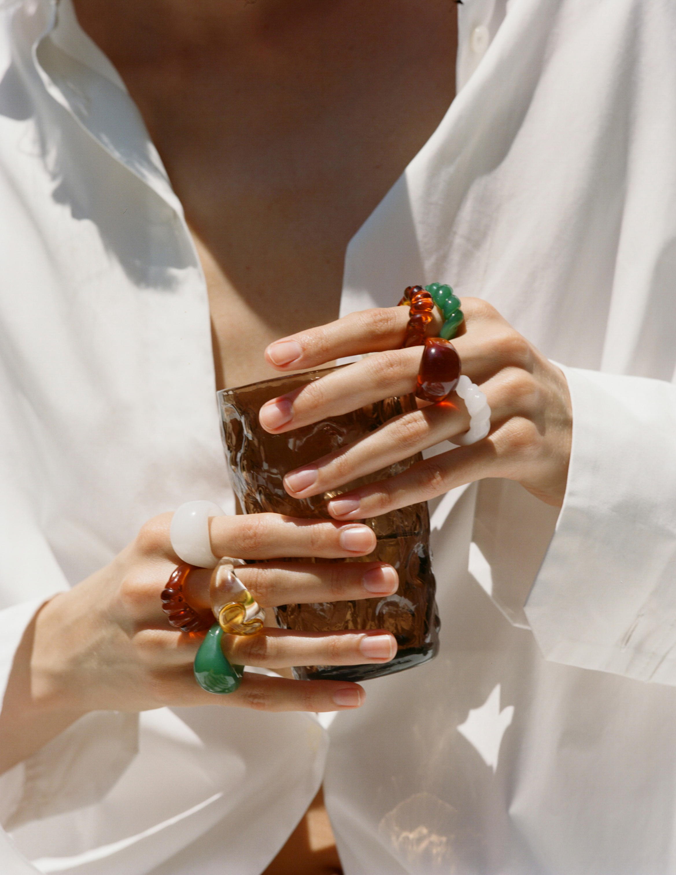 Close-up of hands wearing colorful Pamela Love rings while holding a textured glass, styled with a white shirt