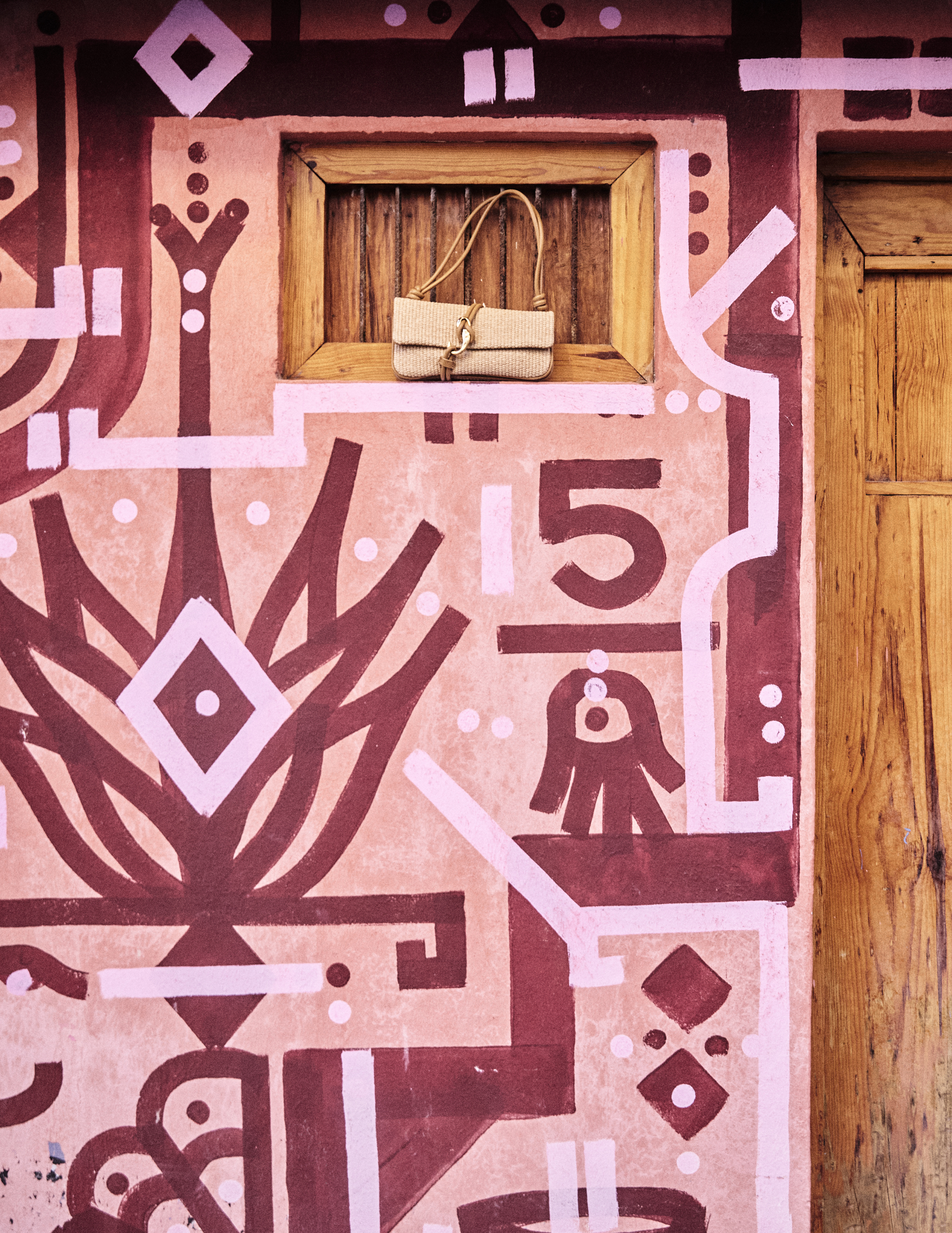 A Dolce Vita neutral woven handbag displayed in a small wooden window, framed by a pink and burgundy geometric mural on an adobe-style wall
