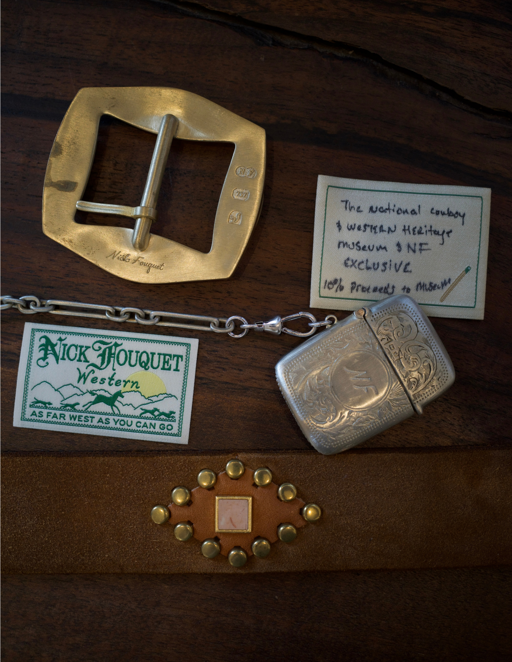 A selection of Nick Fouquet accessories, including a brass belt buckle, engraved silver pieces, and a leather belt, displayed on a wooden surface