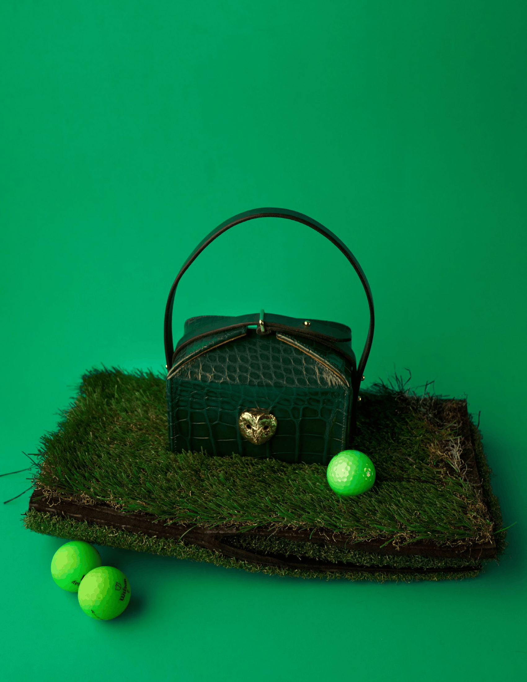 Dark green crocodile-embossed Sydnie Banks handbag with a top handle and gold heart-shaped clasp displayed on a patch of artificial grass, surrounded by bright green golf balls against a matching green backdrop. The editorial still life emphasizes rich color and playful styling.