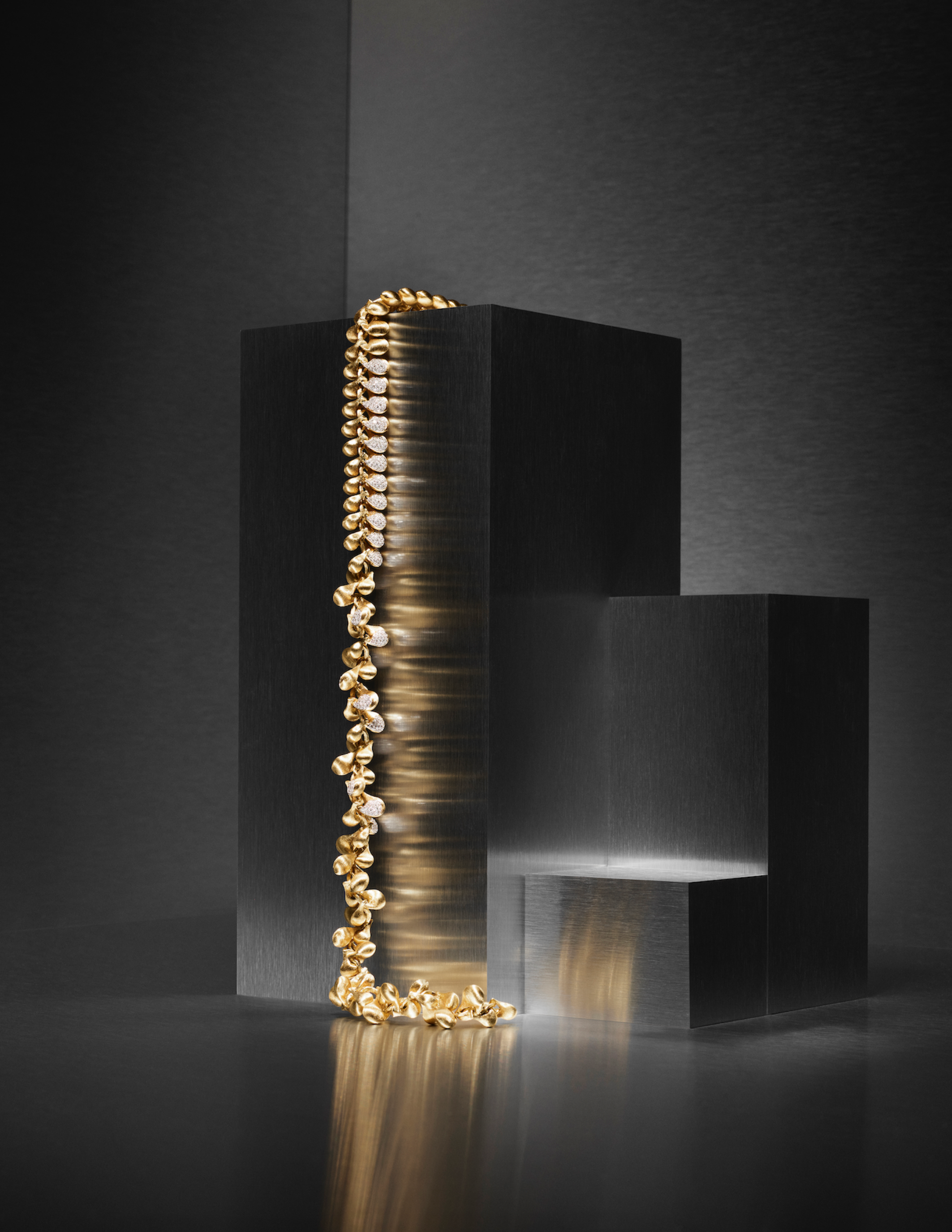 Studio product shot of stacked gold jewelry pieces displayed on dark geometric pedestals.