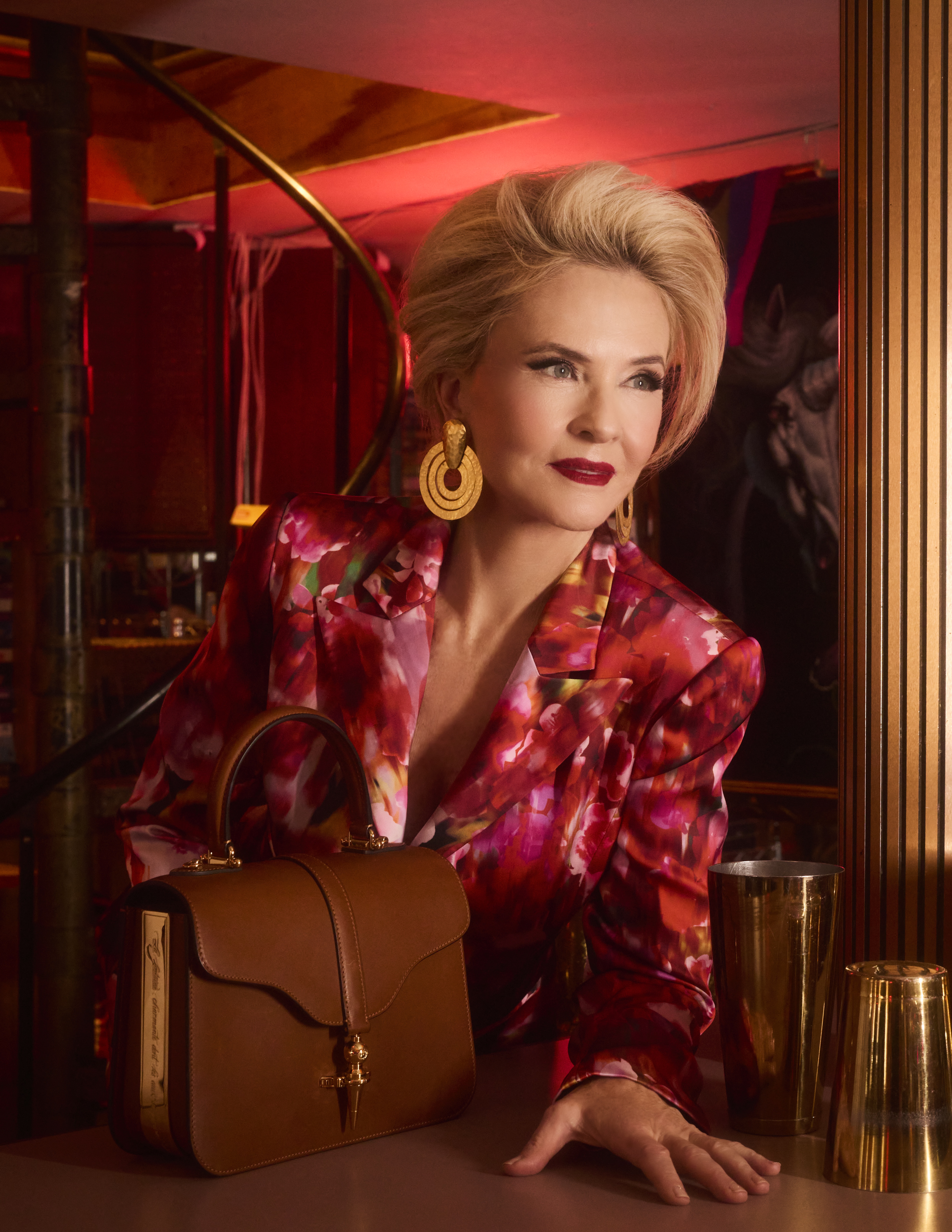 Cornelia Guest leans against a bar in a red floral blazer, wearing bold gold earrings and holding a structured brown leather handbag by Joseph Duclos, posed against a richly lit, retro interior.