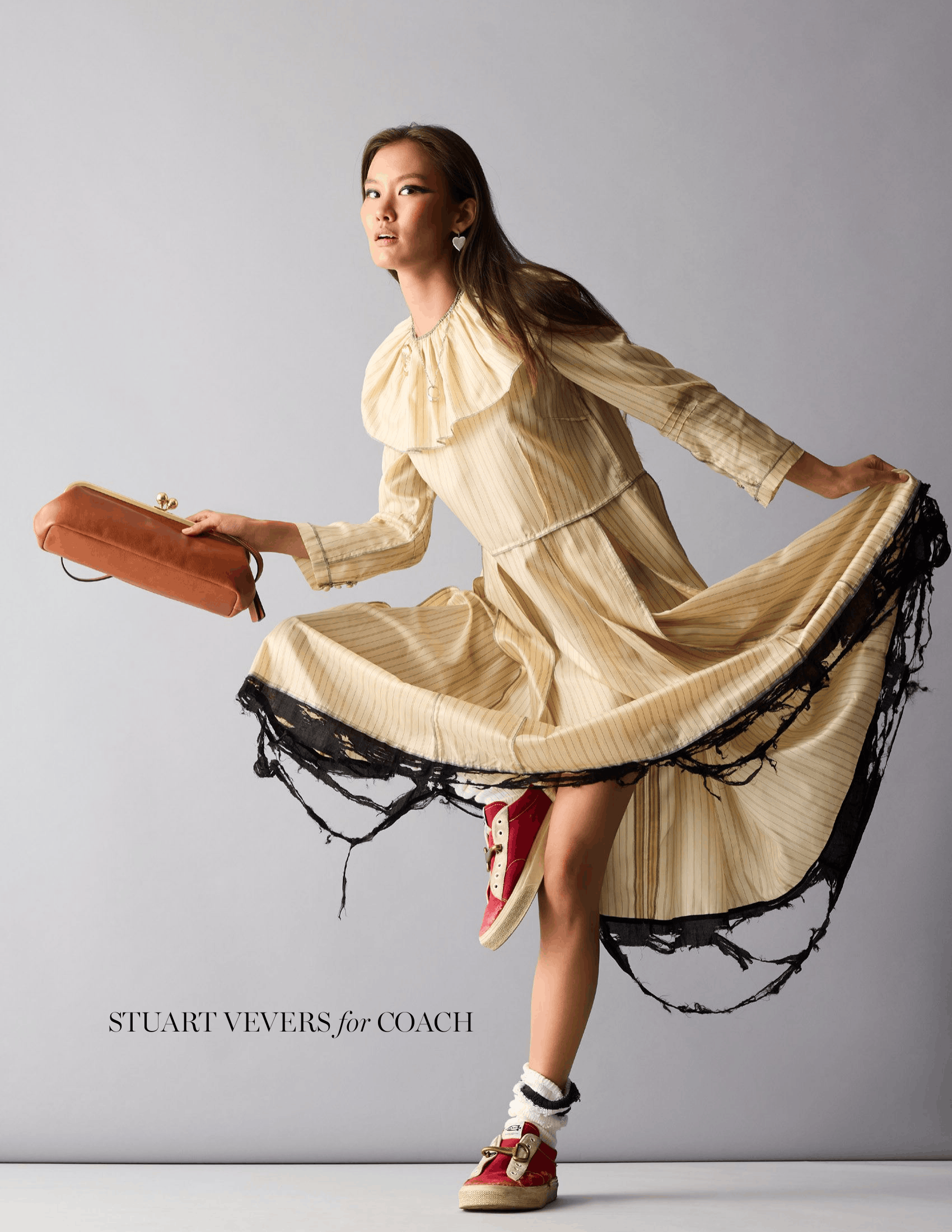 Model Almina Samtonova in a flowing cream-and-beige pleated dress with dramatic black fringe trim, holding a structured brown clutch and wearing red sneakers. Text at the bottom reads “STUART VEVERS for COACH.”