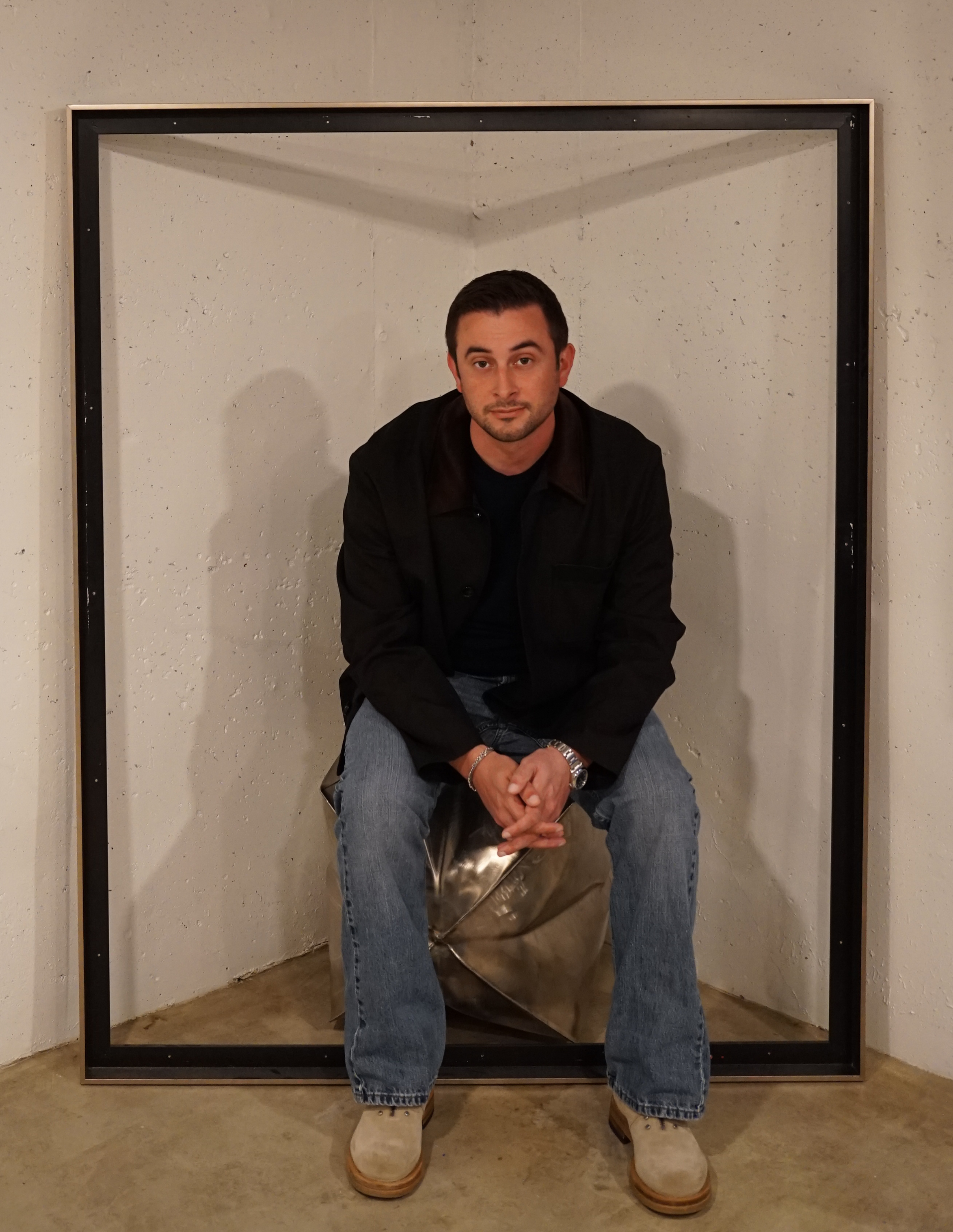 Designer Brady Legler seated inside a large black rectangular frame in his studio, wearing a black jacket, jeans, and tan shoes