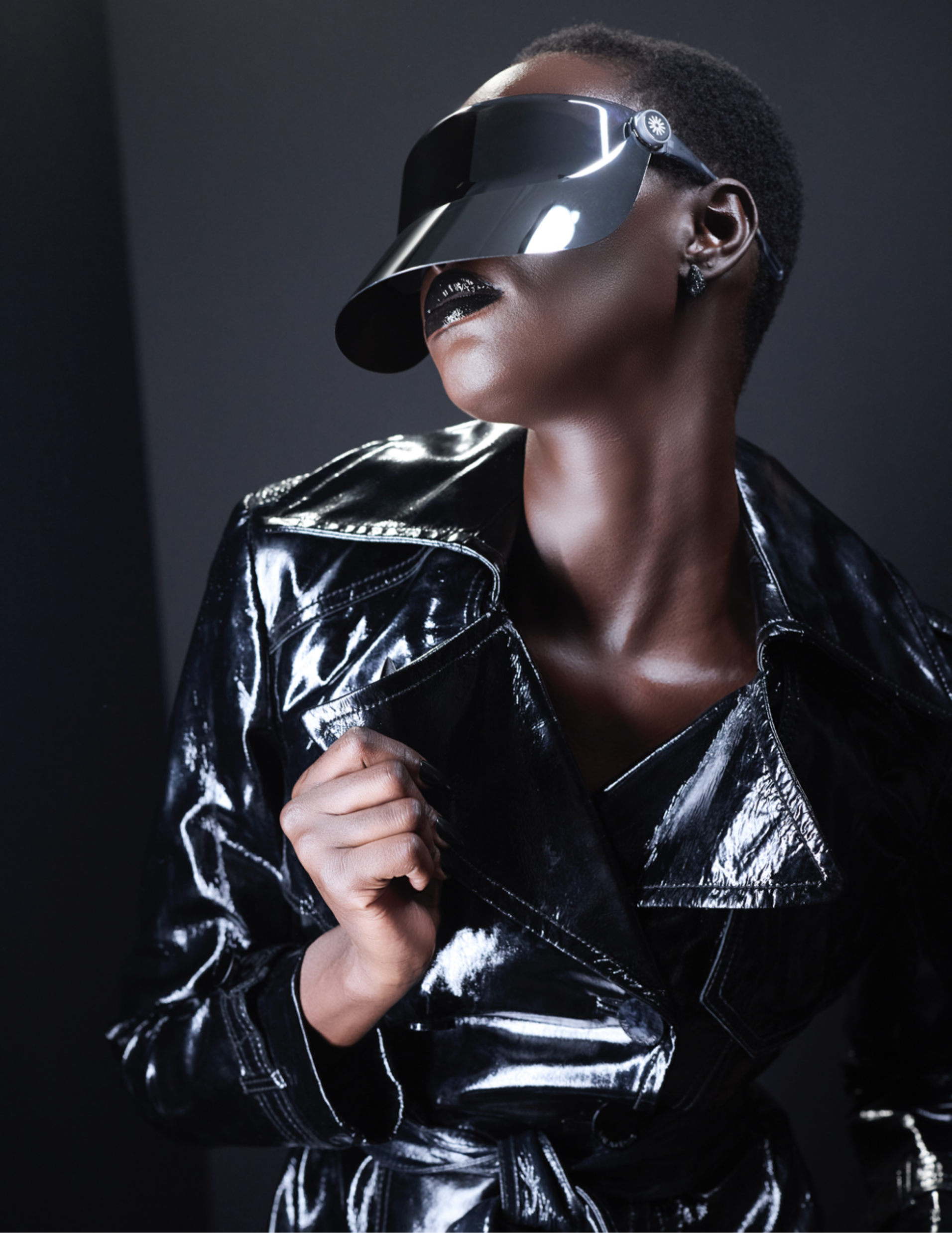 Model Molie Onyongo wears a glossy black trench coat and futuristic mirrored visor by SOUL-A with black statement earrings from Brosway Italia and shimmering black lipstick against a dark studio backdrop.