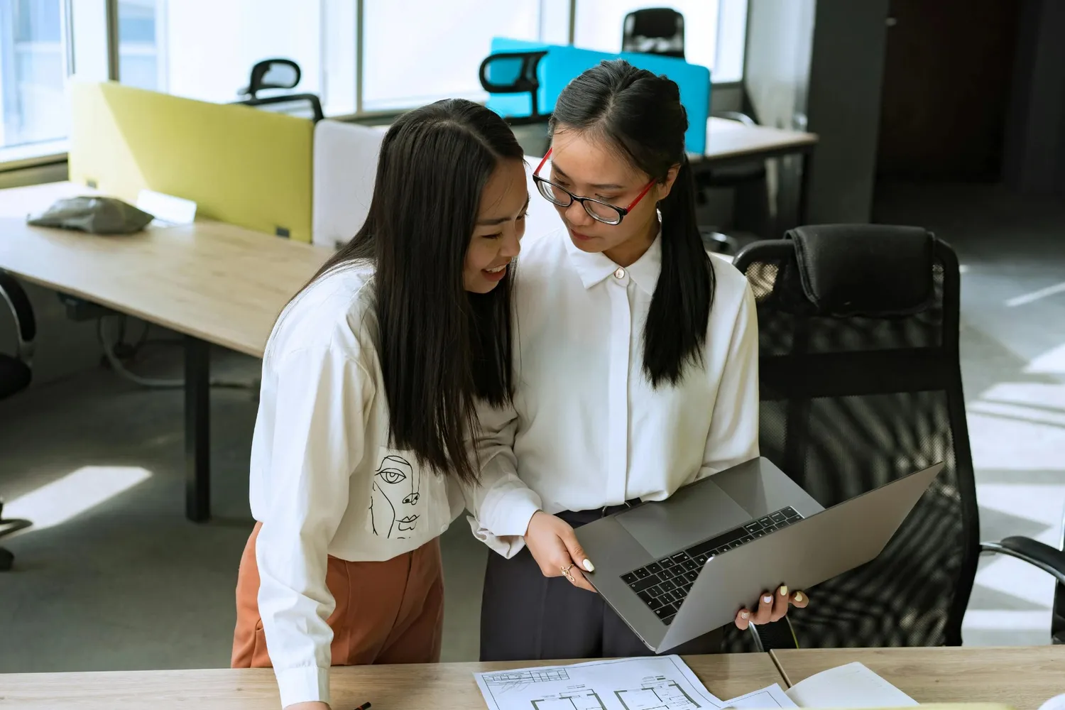 Two women working together in an office, collaborating on thesis editing and proofreading while reviewing documents and discussing on a laptop.