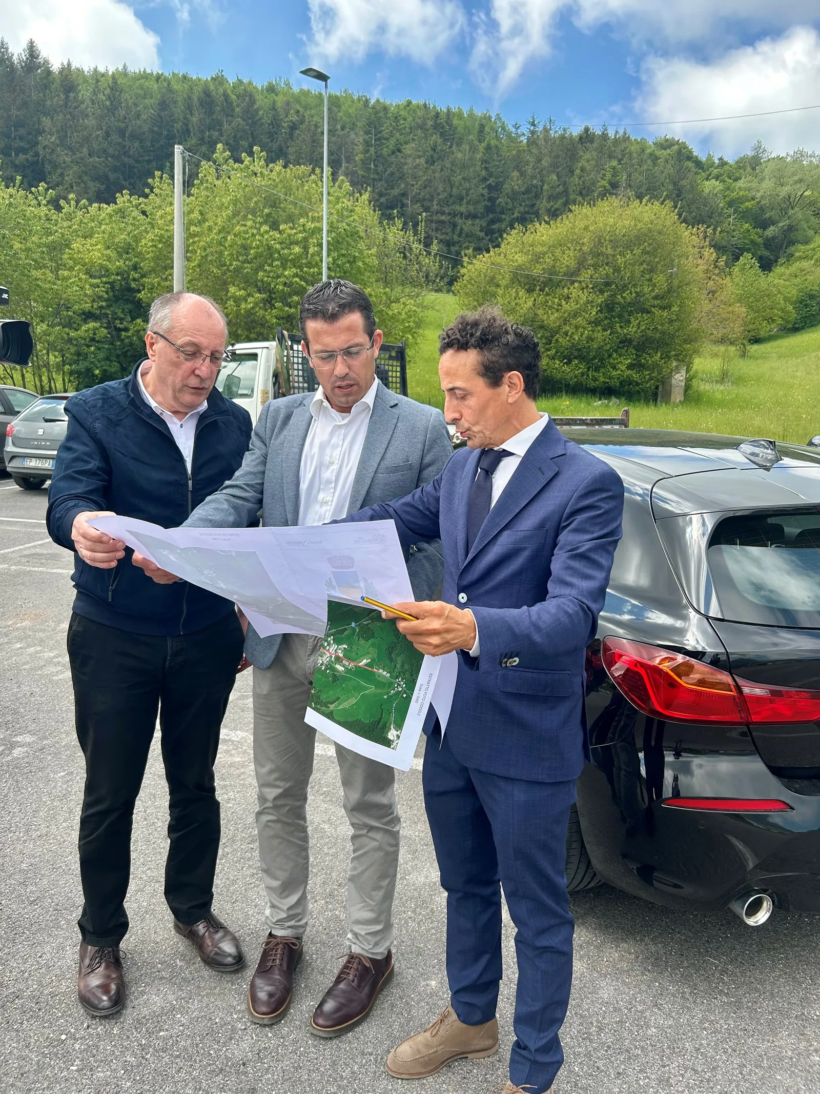 Pianificazione all'aperto: tre esperti analizzano mappe e progetti in un paesaggio verde e soleggiato.