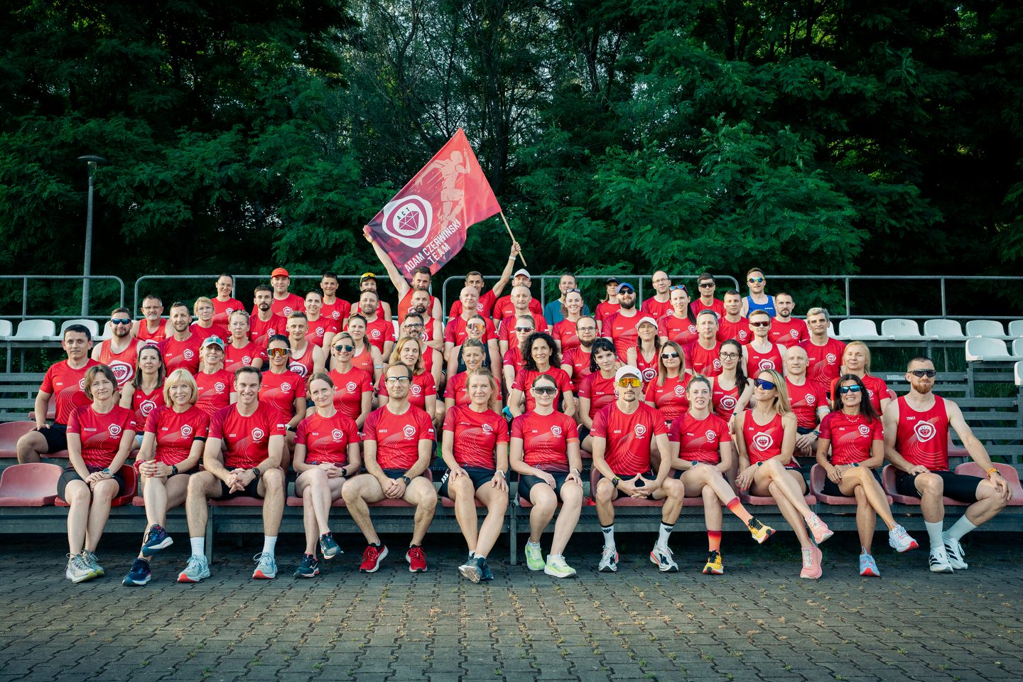 Drużyna #AdamCzerwińskiTeam w czerwonych strojach pozuje na trybunach stadionu z klubową flagą
