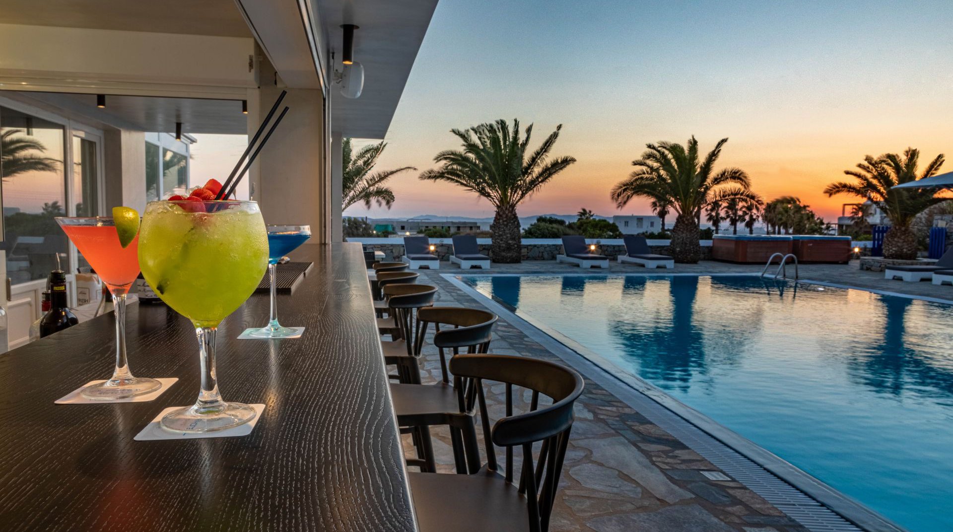 The pool bar of the Daylight Wing during the sunset with the cocktails, the pool, and the sunbeds with the palm trees