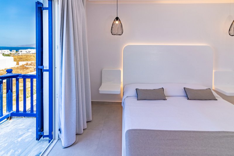 The interior of the room with the side of the bed, the bedside tables, and a glimpse of the island and the sea from the balcony