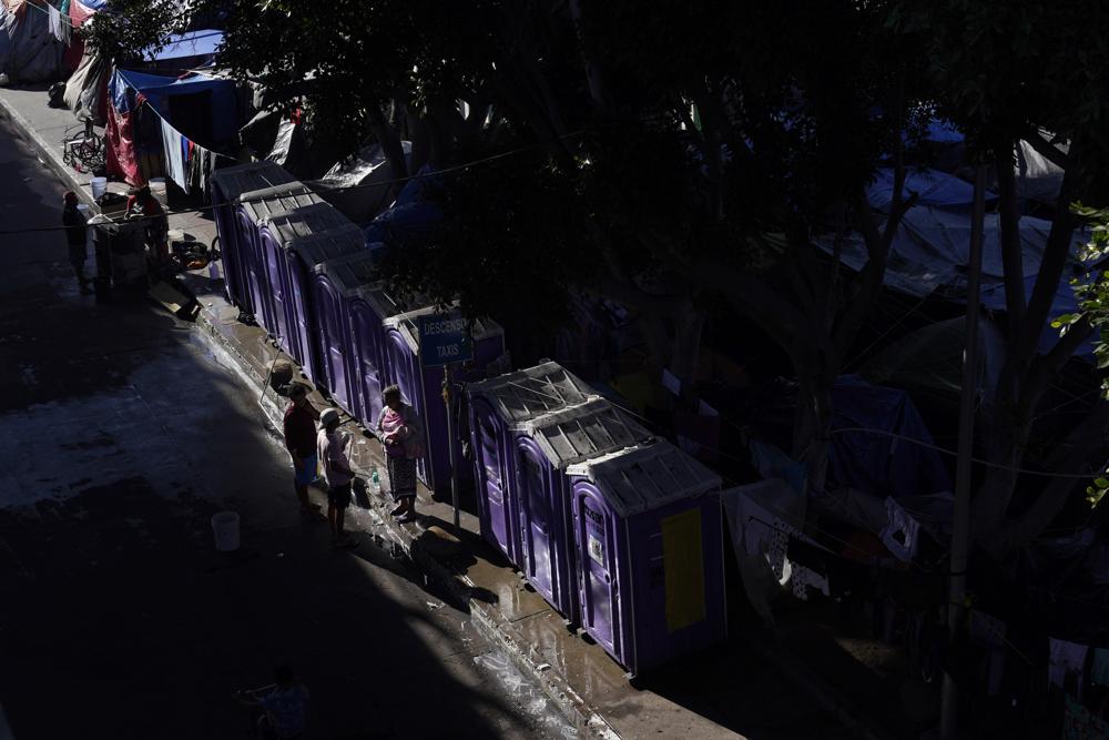 Migrantes charlan enfrente de los baños portátiles en un asentamiento que bloquea la entrada a un cruce peatonal a Estados Unidos (arriba), el 8 de noviembre de 2021, en Tijuana, México. El campamento, ahora rodeado por una cerca metálica tras una operación policial el 28 de octubre, sirve de alojamiento temporal a cientos de migrantes que esperan solicitar asilo en Estados Unidos. (AP Foto/Gregory Bull)