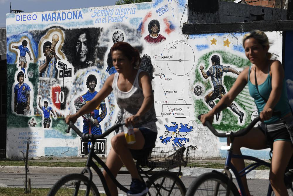 Dos ciclistas pasan junto a un mural que muestra a Diego Maradona con las distintas camisetas que lució durante su carrera en el barrio bonaerense de Lomas de Marilo el 22 de noviembre del 2201. El 25 de noviembre se cumple el primer aniversario de la muerte de Maradona . (Foto AP / Gustavo Garello)