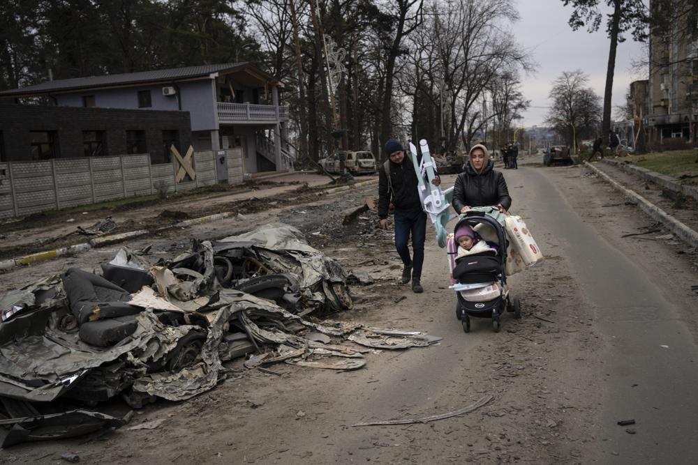 Una familia pasa junto a un automóvil aplastado por un tanque ruso en Bucha, en las afueras de Kiev, Ucrania, el martes 5 de abril de 2022. El presidente de Ucrania planeó dirigirse al organismo más poderoso de la ONU el martes después de que surgieron pruebas aún más espeluznantes de masacres de civiles. en áreas de las que las fuerzas rusas se retiraron recientemente.  (Foto AP/Rodrigo Abd)