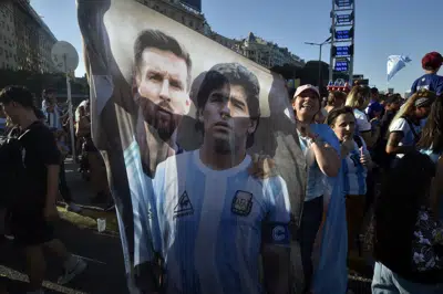 Una bandera con la imagen de Lionel Messi (izquierda) y el extinto astro Diego Maradona mientras aficionados observan la victoria de Argentina ante Croacia en las semifinales del Mundial, el martes 13 de diciembre de 2022, en Buenos Aires. (AP Foto/Gustavo Garello)