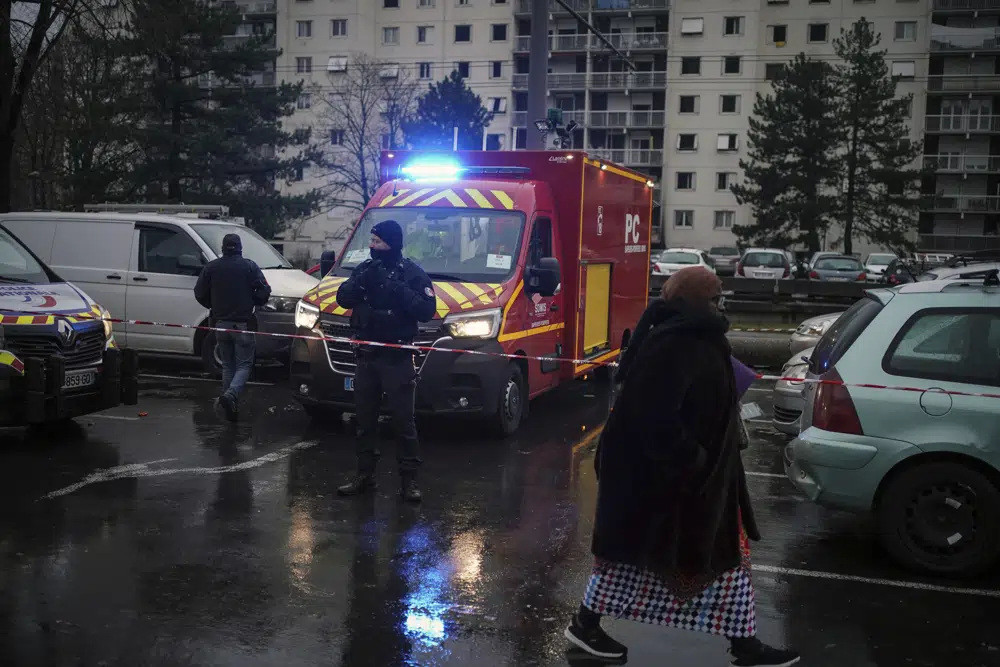 Police officer stands by a firefighter truck next to apartment buildings seen in background at Le Mas du Taureaum neighborhood, in Vauls en Velin, outside Lyon, central France, Friday, Dec. 16, 2022. French authorities say 10 people including five children died in a fire in an apartment building outside the city of Lyon. The cause of the fire is being investigated. (AP Photo/Laurent Cipriani)