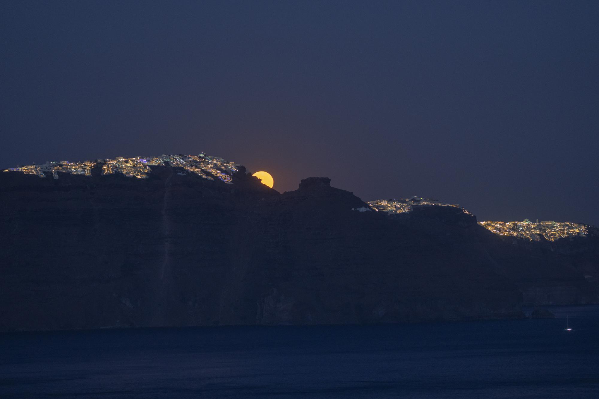 La luna sale detrás del pueblo de Imerovigli, a la izquierda, y el promontorio rocoso de Skaros, a la derecha, en la isla griega de Santorini el martes 14 de junio de 2022. El Monasterio Católico de Santa Catalina fue fundado en Skaros en 1596, pero después un terremoto se trasladó a la ciudad de Thira, extremo derecho.  (Foto AP/Petros Giannakouris)