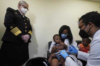 La pediatra Emy Jean-Marie, en el centro, sostiene a su hijo de nueve meses, Adedeji Adebayo, junto a su hija de tres años, Emiola Adebayo, mientras el doctor Nizar Dowla, a la derecha, administra una vacuna ante la mirada de la almirante Rachel Levine, secretaria asistente del Departamento de Salud y Servicios Humanos, el 28 de junio de 2022.   en el centro de salud Borinquen en Miami, Florida. (AP Foto/Wilfredo Lee)