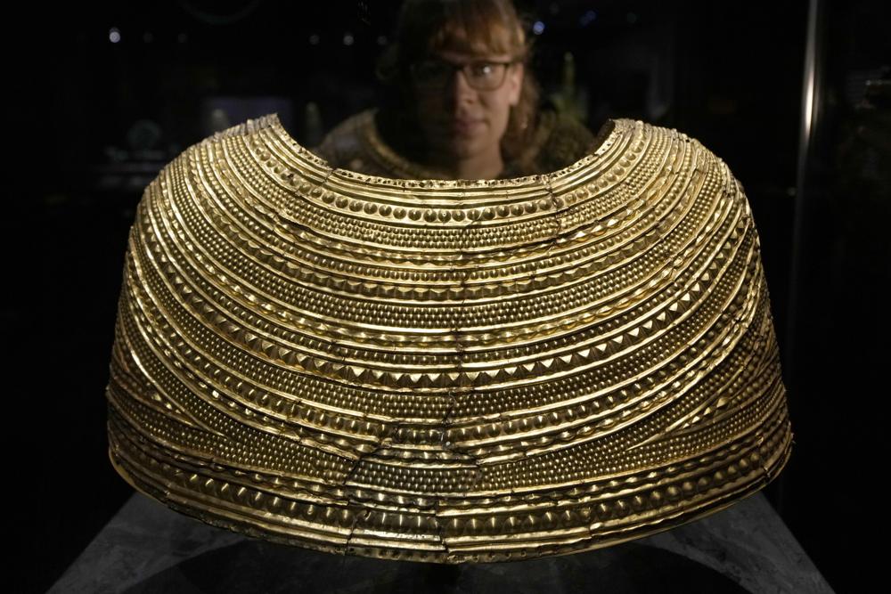 Una miembro del personal posa junto a una capa de oro de entre 1600 y 1900 a.C., hallada en Mold, en Gales, en la exposición "The World of Stonehenge" en el Museo Británico de Londres el lunes 14 de febrero de 2022. (Foto AP/Alastair Grant)