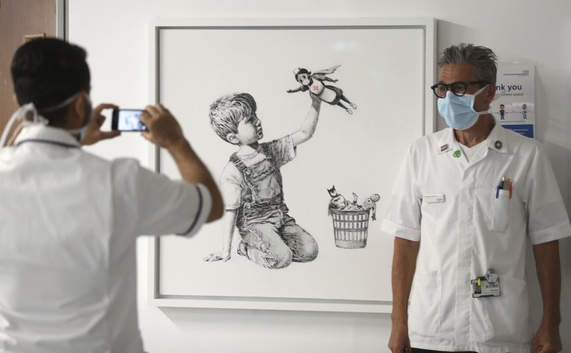 Un membre del personal sanitari fotografia un company, amb bata blanca, mascareta i ulleres, al costat d’un quadre de Banksy, emmarcat, penjat en una paret, que representa un xiquet jugant amb la nina d’una superheroïna infermera.