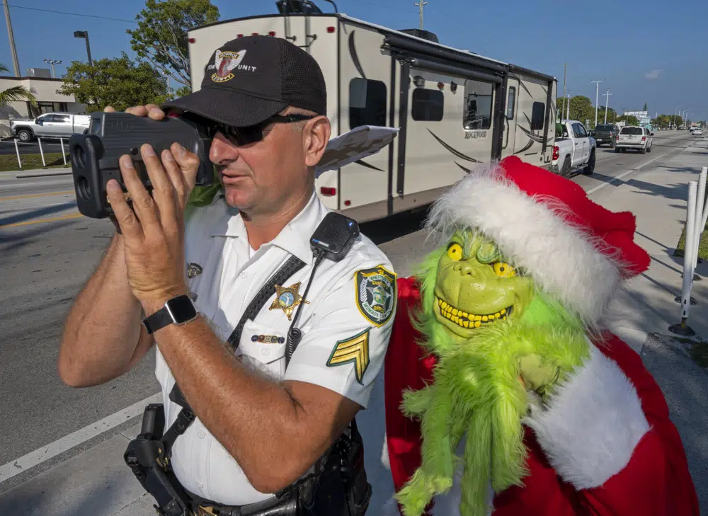 In this photo provided by the Florida Keys News Bureau, Monroe County Sheriff's Office Colonel Lou Caputo, right, costumed as the Grinch, watches Sgt. Greg Korzan, left, as he uses a laser speed detector to check speeds of motorists traveling through a school zone on the Florida Keys Overseas Highway Tuesday, Dec. 13, 2022, in Marathon, Fla. For drivers slightly speeding through the area, Caputo offers them the choice between an onion or a traffic citation. (Andy Newman/Florida Keys News Bureau via AP)