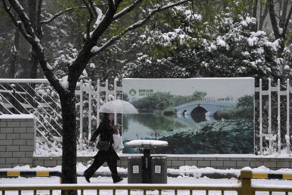 Una persona pasa junto a un cartel de un parque con una fotografía que muestra el lugar en verano, durante una nevada en Beijing, China, el domingo 7 de noviembre de 2021. (AP Foto/Ng Han Guan)