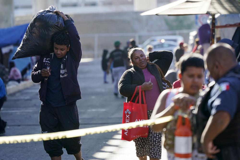 Migrantes cargan con sus pertenencias en un asentamiento improvisado que bloquea la entrada a un cruce peatonal a Estados Unidos (arriba), el 8 de noviembre de 2021, en Tijuana, México. El campamento, ahora rodeado por una cerca metálica tras una operación policial el 28 de octubre, sirve de alojamiento temporal a cientos de migrantes que esperan solicitar asilo en Estados Unidos. (AP Foto/Gregory Bull)