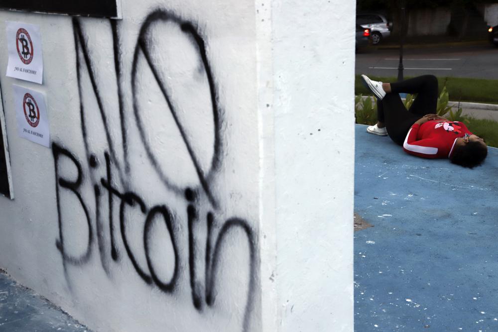 Una mujer descansa cerca de una pared pintada con aerosol con un mensaje de "No Bitcoin" durante una protesta contra el gobierno en San Salvador, El Salvador, el miércoles 1 de septiembre de 2021. (AP Foto/Salvador Meléndez)