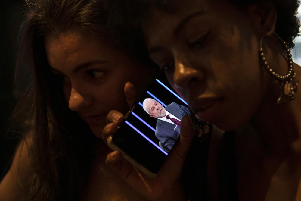 FILE - Women listen on a mobile phone to an interview of former president Luiz Inacio Lula da Silva, who is running again for president, with Jornal Nacional on TV Globo, outside a bar in Rio de Janeiro, Brazil, Thursday, August 25, 2022. The Superior Electoral Court, the country's top electoral authority, announced Thursday, Oct, 20, that it would be banning 