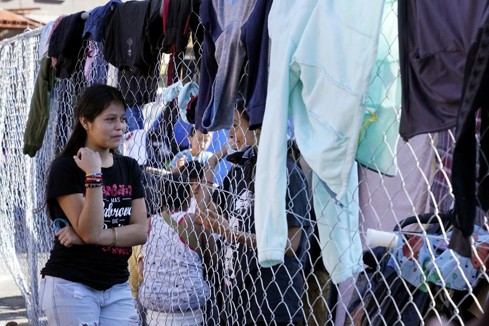 Migrantes hablan a través de la cerca de un asentamiento improvisado que bloquea la entrada a un cruce peatonal a Estados Unidos (arriba), el 8 de noviembre de 2021, en Tijuana, México. El campamento, ahora rodeado por una cerca metálica tras una operación policial el 28 de octubre, sirve de alojamiento temporal a cientos de migrantes que esperan solicitar asilo en Estados Unidos. (AP Foto/Gregory Bull)