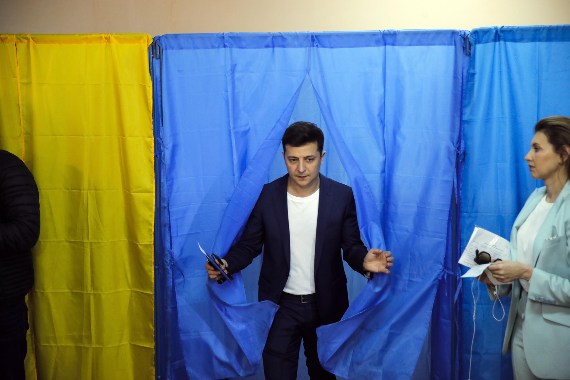 ФАЙЛ — Украинский комик и кандидат в президенты Владимир Зеленский (в центре) выходит из кабинки на избирательном участке, пока его жена Елена Зеленская ждет во время второго тура президентских выборов в Киеве, Украина, воскресенье, 21 апреля 2019 года. Год назад, с русским силы, надвигающиеся на столицу Украины, западные лидеры опасались за жизнь президента Владимира Зеленского, а США предложили ему путь к отступлению. Зеленский отказался, заявив о своем намерении остаться и защищать независимость Украины. (AP Photo/Вадим Гирда, файл)