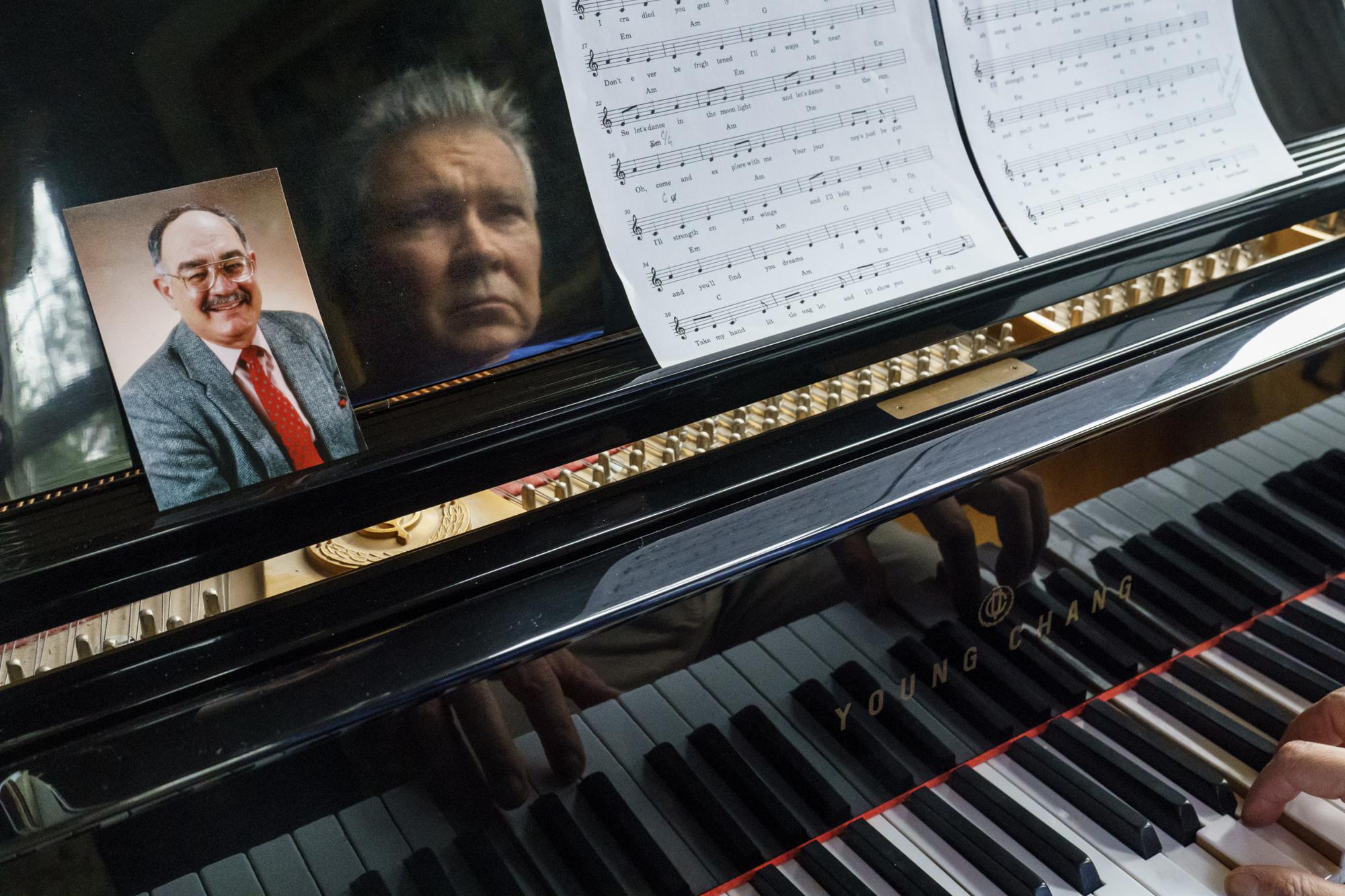 A photo of Neil Lawyer sits on the piano as his son, David Lawyer, plays in his home in Bellevue, Wash., Sunday, March 20, 2022. When the elder Lawyer died of complications from COVID-19 on March 8, 2020, the U.S. toll stood at 30. At weddings, he joined his sons to serenade brides and grooms in a makeshift ensemble dubbed the Moose-Tones. Last October, when one of his granddaughters married, it marked the first family affair without Lawyer there to hold court. The Moose-Tones went on without him. "He would have just been beaming because, you know, it was the most important thing in the world to him late in life, to get together with family," David says. "I can honestly tell you he was terribly missed." (AP Photo/David Goldman)