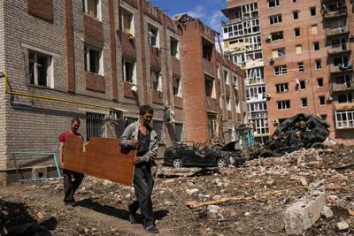 Dos hombres llevan un panel arbolado junto a edificios muy dañados y automóviles destruidos en un bombardeo ruso en Bakhmut, al este de Ucrania, el martes 24 de mayo de 2022. La ciudad de Bakhmut ha estado siendo objeto de crecientes ataques de artillería, particularmente durante la última semana, a medida que las fuerzas rusas intentan avanzar para rodear la ciudad de Sieverodonetsk al noreste. (AP Foto/Francisco Seco)