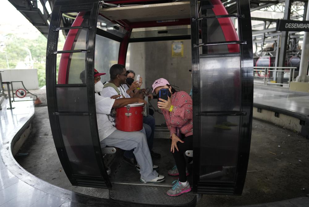 Una turista se toma una foto mientras abordan un teleférico acompañado por un músico local durante un recorrido organizado por "Cumbe Tour" al barrio San Agustín de Caracas, Venezuela, el sábado 21 de agosto de 2021. Cumbe Tour trae a turistas, tanto nacionales como extranjeros, a San Agustín para mostrarles cómo viven los venezolanos de bajos ingresos en lo que solía ser en otrora uno de los barrios más peligrosos de la ciudad. (Foto AP/Ariana Cubillos)