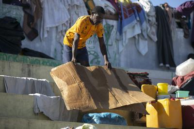 Un hombre seca un pedazo de cartón que utiliza para dormir en la plaza pública Hugo Chávez, convertida en un refugio para familias obligadas a huir de sus casas debido a los enfrentamientos entre pandillas, el jueves 20 de octubre de 2022, en Puerto Príncipe, Haití. (AP Foto/Odelyn Joseph)