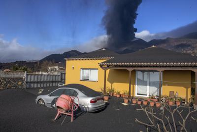 Una capa de ceniza volcánica cubre una casa, un automóvil y sus alrededores mientras en el fondo un volcán sigue en erupción en la isla canaria de La Palma, España, el lunes 4 de octubre de 2021. (AP Foto/Saul Santos)