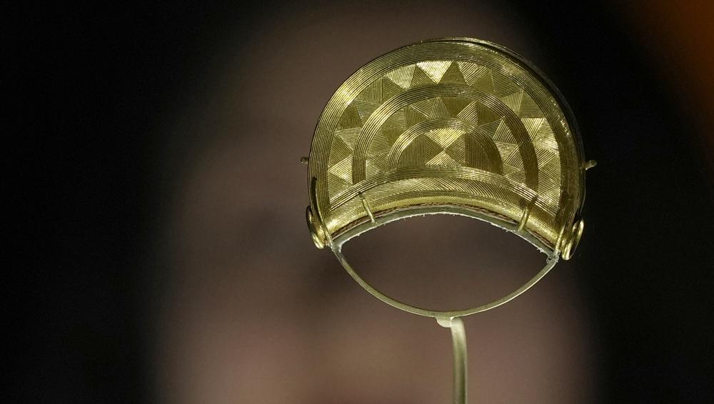 Una miembro del personal posa junto a un broche de oro de Shropshire, Inglaterra, del año 1000 AEC, en la exposición "The World of Stonehenge" en el Museo Británico de Londres, el lunes 14 de febrero de 2022. (Foto AP/Alastair Grant)