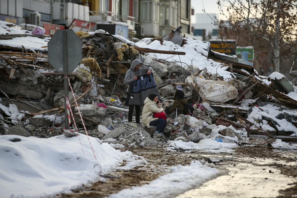 People sit by a collapsed building in Malatya, Turkey, Tuesday, Feb. 7, 2023. Search teams and aid are pouring into Turkey and Syria as rescuers working in freezing temperatures dig through the remains of buildings flattened by a magnitude 7.8 earthquake. (AP Photo/Emrah Gurel)