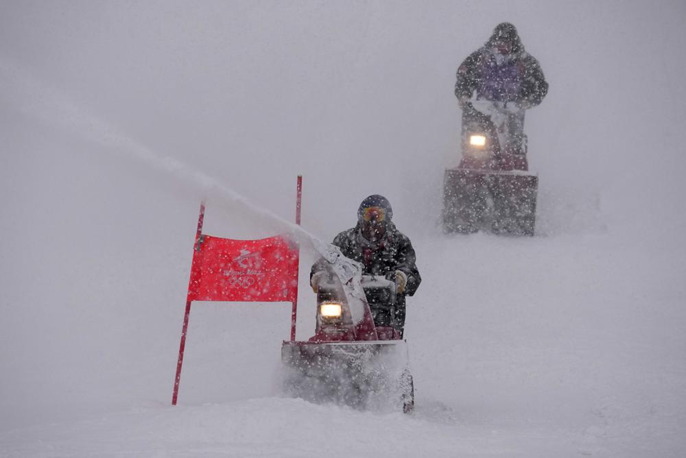 Trabajadores de la pista soplan la nieve de la pista de slalom gigante masculino mientras la nieve cae en la sede de esquí alpino de los Juegos Olímpicos de Invierno de 2022, el domingo 13 de febrero de 2022, en el distrito de Yanqing de Beijing.  (Foto AP/Robert F. Bukaty)