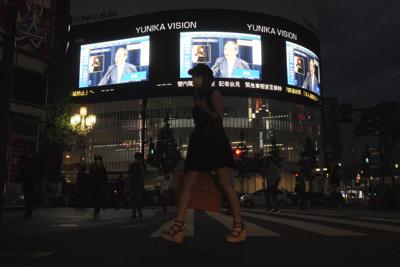 Varias personas caminan por una calle de Tokio mientras al fondo de observan pantallas con la imagen del primer ministro japonés, Yoshihide Suga, hablando en vivo durante una conferencia de prensa sobre la respuesta de Japón a la pandemia de coronavirus, la noche del jueves 17 de junio de 2021. (AP Foto/Eugene Hoshiko)