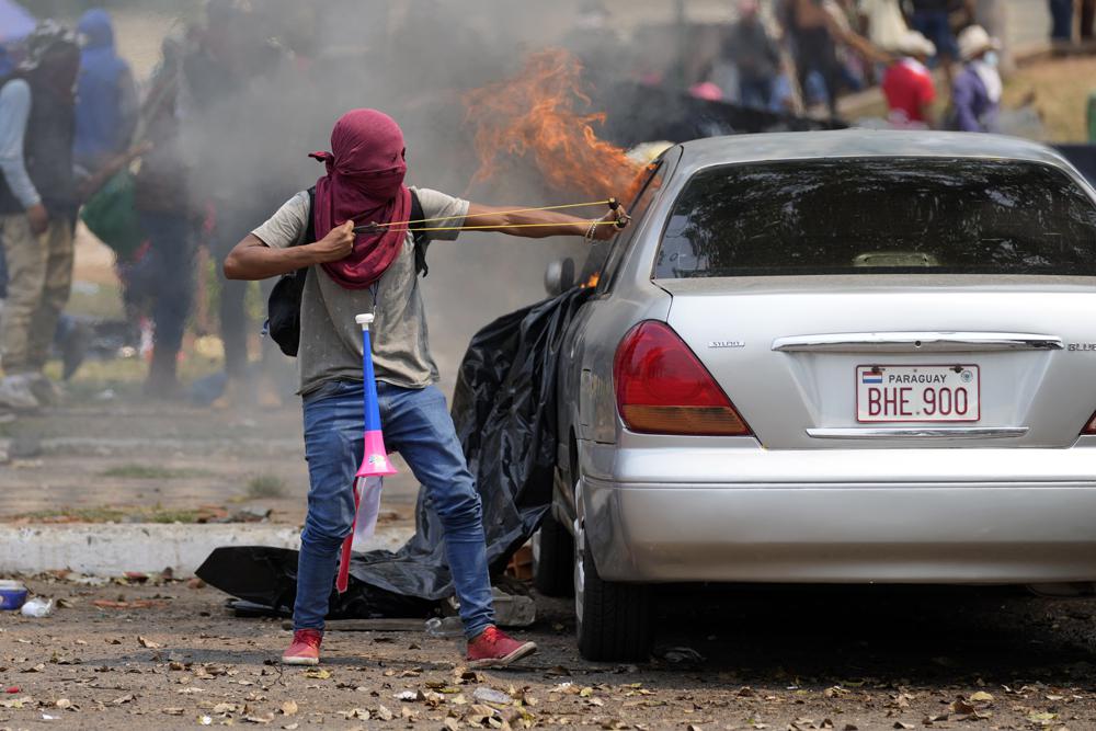 A masked protester uses a slingshot during clashes with police during an Indigenous protest against a proposed bill that criminalizes land invasions, outside Congress in Asuncion, Paraguay, Wednesday, Sept. 29, 2021. If the bill passes it would affect several Indigenous communities who reside on improvised settlements pending the restitution of their lands. (AP Photo/Jorge Saenz)