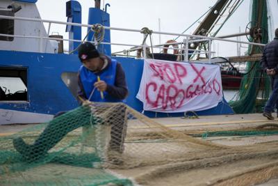 Un pescador repara una red frente a su barco pesquero con una pancarta que dice "alto para aumentar la gasolina", en el puerto romano de Fiumicino, el viernes 11 de marzo de 2022. Los pescadores, que se enfrentaban a enormes picos en los precios del petróleo, se quedaron en el puerto, reparando las redes en lugar de lanzarlas. En ninguna parte más que en Italia, la tercera economía más grande de la Unión Europea, la dependencia de la energía rusa está cobrando un mayor precio en la industria. (AP Foto/Andrew Medichini)
