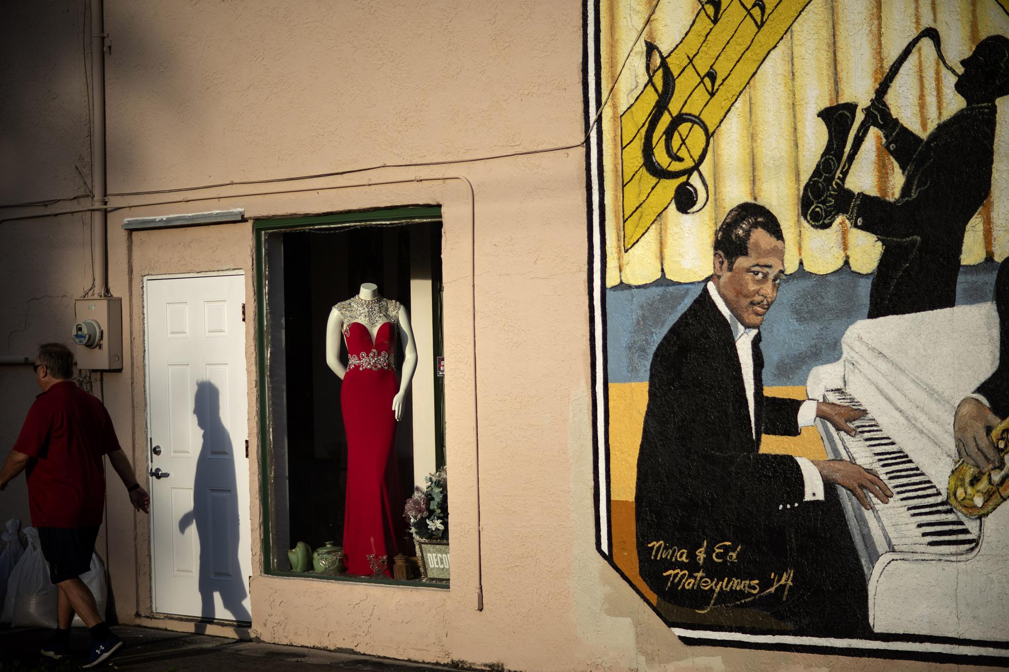 An evening gown is displayed in a store window next to a mural titled "Harlem Nights in Palatka," featuring musicians from the town that formed a 1920s jazz band, as a pedestrian walks by in downtown Palatka, Fla., on Tuesday, April 13, 2021. (AP Photo/David Goldman)