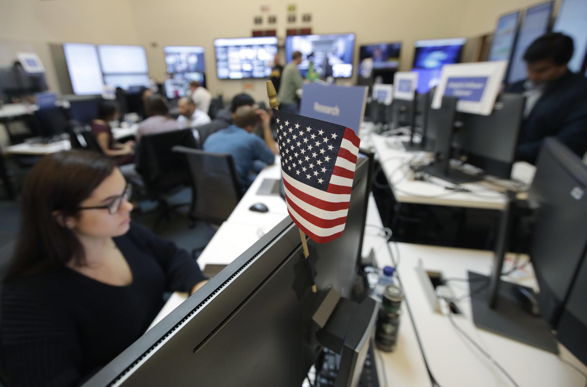 FILE – In this Oct. 17, 2018, file photo, a flag of the United State is shown between monitors as workers sit at their desks during a demonstration in the war room, where Facebook monitors election related content on the platform, in Menlo Park, Calif. From complaints whistleblower Frances Haugen has filed with the SEC, along with redacted internal documents obtained by The Associated Press, the picture of the mighty Facebook that emerges is of a troubled, internally conflicted company, where data on the harms it causes is abundant, but solutions are halting at best. (AP Photo/Jeff Chiu, File)