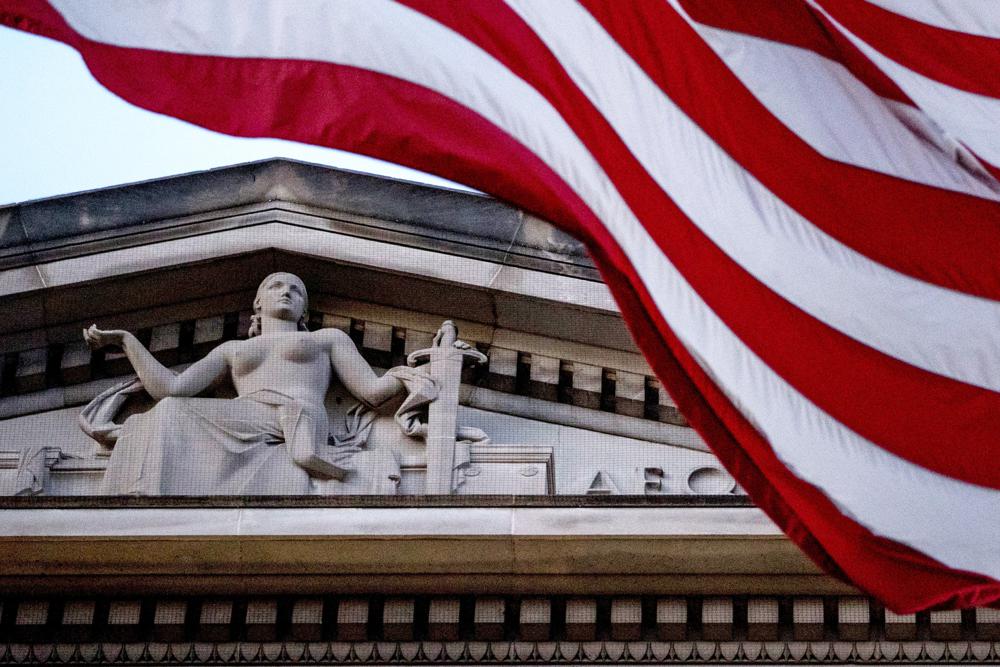 FILE - An American flag flies outside the Department of Justice in Washington, March 22, 2019. The Justice Department will begin transferring thousands of inmates out of federal prisons across the U.S. this week as it implements part of the First Step Act. It is part of a sweeping criminal justice measure signed into law in 2018. The Justice Department is publishing a rule in the Federal Register on Thursday that codifies how it will implement the program for earned time credits. (AP Photo/Andrew Harnik, File)