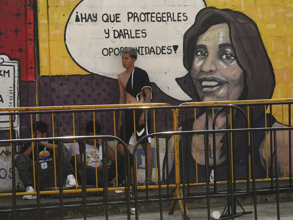 Migrantes esperan su turno para regularizar su situación migratoria junto a un mural pintado frente al Instituto Nacional de Migración en Tapachula, estado de Chiapas, México, el martes 4 de octubre de 2022. Los procesos de regulación migratoria puede llevar meses y cualquier oferta para agilizar esos trámites es tentadora para quien no quiere esperar ni tampoco arriesgar su vida cruzando la frontera hacinado en un camión. (AP Foto/Marco Ugarte)