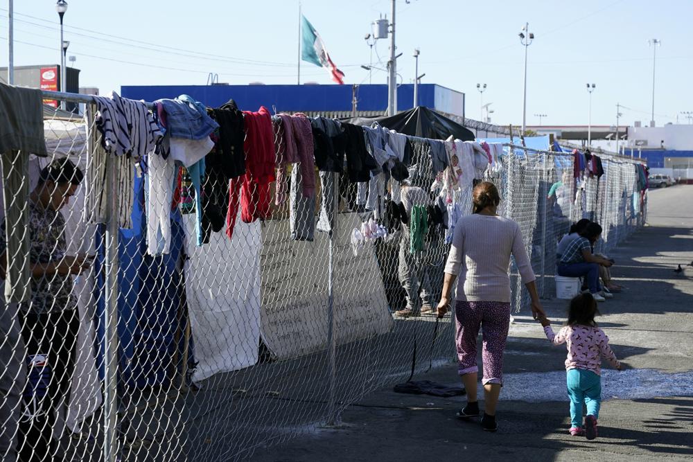 Migrantes caminan junto a la cerca de un campamento improvisado que bloquea la entrada a un cruce peatonal a Estados Unidos (arriba), el 8 de noviembre de 2021, en Tijuana, México. El campamento, ahora rodeado por una cerca metálica tras una operación policial el 28 de octubre, sirve de alojamiento temporal a cientos de migrantes que esperan solicitar asilo en Estados Unidos. (AP Foto/Gregory Bull)