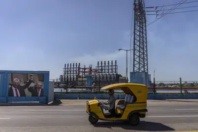 Un coco taxi pasa frente a una fotografía del expresidente cubano Raúl Castro y el actual presidente Miguel Díaz-Canel y un barco de suministro de energía de Turquía que alimenta la ciudad de La Habana, Cuba, el martes 18 de abril de 2023. (AP Foto/Ramón Espinosa)