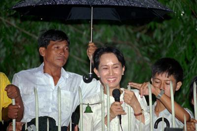 FILE - An aid holds an umbrella over Burmese democratic leader Aung San Suu Kyi during her daily appearance at the gate of her Yangon (formerly known as Rangoon) home from where she greets a crowd of supporters that gathered in the street on July 16, 1995. A court in Myanmar sentenced the country’s ousted leader, Aung San Suu Kyi, to four years in prison on Monday, Dec. 6, 2021, after finding her guilty of incitement and violating coronavirus restrictions, a legal official said. (AP Photo/Anat Givon, File)