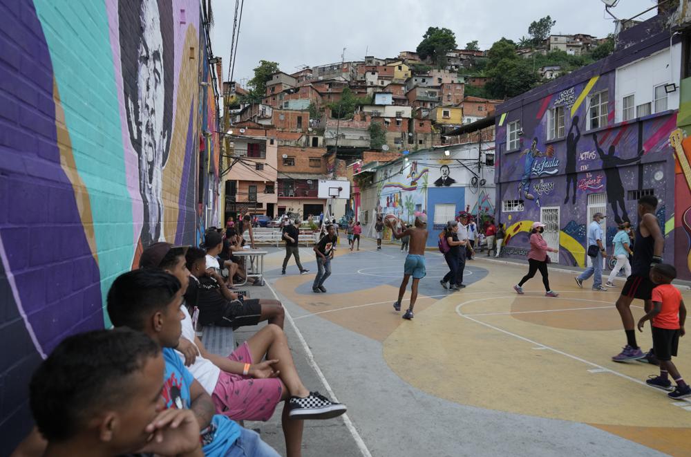 Jóvenes juegan baloncesto mientras un grupo de turistas camina por la cancha durante un recorrido por el barrio de San Agustín, organizado por Cumbe Tour en Caracas, Venezuela, el sábado 21 de agosto de 2021. Cumbe Tour trae a turistas, tanto extranjeros como nacionales, a San Agustín para mostrarles cómo viven los venezolanos de menores ingresos en lo que solía ser uno de los barrios más peligrosos de la ciudad. (Foto AP/Ariana Cubillos)